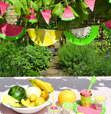 Gebastelte Sommergirlande aus Papptellern, die wie Melonen angemalt sind. Im Vordergrund sieht man eine gedeckten Tisch mit Melonen und Früchten.