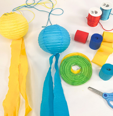 Lampions mit Krepppapier geschmückt und daneben all die benötogten Materialien: Lampion-Rohlinge, Krepppapierrollen, Schnur und eine Schere.