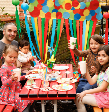 Vier Kinder und ein Erwachsener sitzen an einem roten Tisch im Freien und lachen in die Kamera. Die Szene ist sommerlich-festlich. Der Hintergrund ist geschmückt mit Luftballons und einer Papiergirlande.
