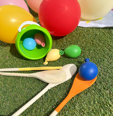Holzlöffel, mit Sand gefüllten Luftballons und bunte Eimer die alle auf einer Wiese liegen.