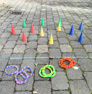 Gebasteltes Wurfspiel: Zu Ringen gedrehte Pfeifenputzer und angemalte Papp-Kegel. Die Ringe liegen vorne auf den Boden, dahinter sieht man die mit Kreide eingezeichnete Startlinie, hinter der die bunten Kegel aufgereiht sind.