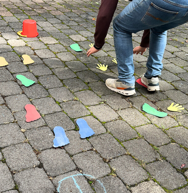 Der Boden wurde zu einem Parkour: Mit Kreide ist die Start- und Ziellinie eingezeichnet. Der Parkour besteht aus Händen und Füßen aus buntem Papier, die auf dem Boden liegen. Auf der echte Seite sieht man einen Mann, der mit seinen Füßen auf ein Paar der Papierfüßen steht und seine Hände auf die Papierhände stützt.