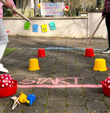 Outdoor-Aufnahme: Auf dem Boden wurde mit Kreide eine Start- und eine Ziellinie gemalt. Mit Eimern wurde ein Parkour für zwei Teilnehmer gebaut. Diese schieben mit einem langen Pappstab jeweils ihren Ball durch den Parkour.