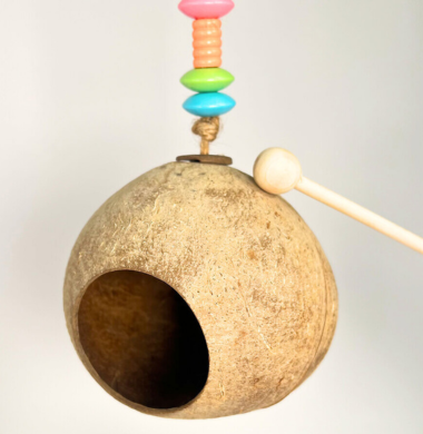 Gebastelte Kokosnuss-Gong aus einer ausgehüllten Kokosnuss, die an einer Schnur hängt, die mit Perlen geschmückt ist. Mit einem Holzstab lässt sich ein Klang erzeugen.