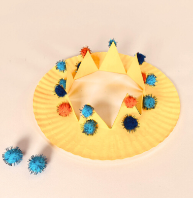 Eine Krone aus einem bemalten Pappteller, der mit Glitzerpompons geschmückt ist.