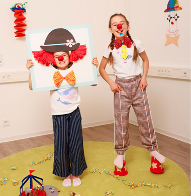 Zwei Mädchen spielen mit gebastelten Clownutensilien. Das eine streckt ihren Kopf durch eine Fotowand, das andere trägt eine bunte Papierkrawatte und steht auf mit Sternen verzierten Laufdollis. Beide tragen rote Clownsnasen und lachen.