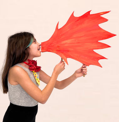 Ein Mädchen häkt eine aus Karton gebastelte Flamme an ihren Mund und tut so, als würde sie Feuer speien.