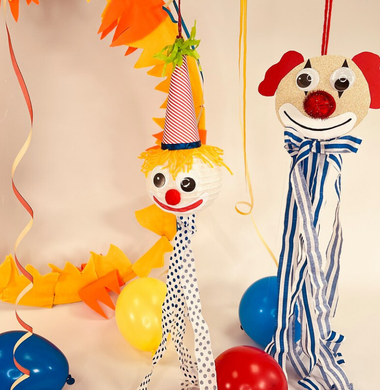 Zwei gebastelte Clowns: einer hat einen Kopf aus einem Lampion, der andere aus Glitzerkarton. Beide haben große Wackelaugen und sind mit Krepppapierstreifen verziert.