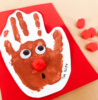 Eine Weihnachtskarte verziert mit einem Handabdruck aus brauner Farbe. Diesem wurden Wackelaugen und eine Pomponnase eaufgeklebt, sodass dieser wie ein Rentier aussieht.