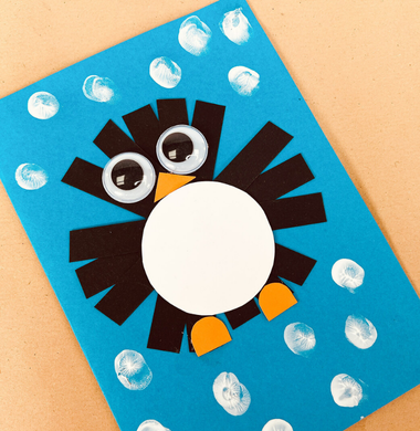 Eine gebastelte Weihnachtskarte aus Fotokarton. Verziert mit Papierstreifen, die im Kreis auf die Karte geklebt wurden und mit Wackelaugen und einem Marker sowie orangenen und weißen Tonpapier, sodass es aussieht wie ein Pinguin.