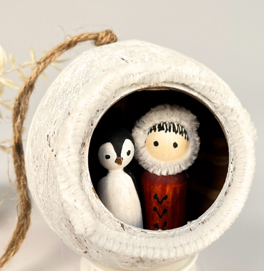 Eine weiß angemalte Kokonuss-Schale, die gefüllt ist mit zwei Holzfiguren. Eine stellt einen Pinguin dar, die andere einen Inuit.