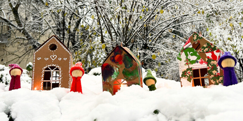 Kleiner Wichtel aus Holzfigur und in Bastelfilz gekleidet steht vor verzierten Papphäusern. Im Hintergrund sieht man Schnee.