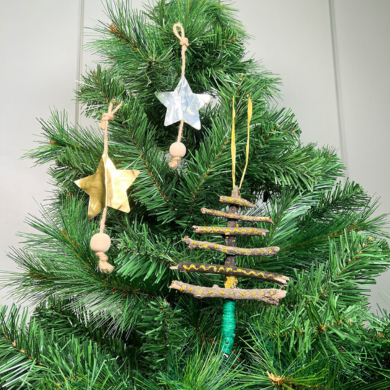 An einem Tannenbaum hängen drei gebastelte Anhänger: zwei sind Sterne aus Glitzerfolie, die an einem Jutegarn hängen und mit einer Holzperle geschmückt sind. Der andere Anhänger besteht aus kleinen Ästen, die in Form eines Tannenbaums zusammengeklebt wurden.