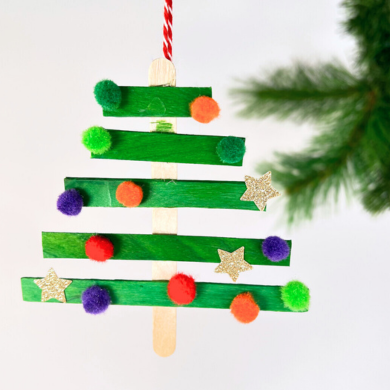 Ein Anhänger in Form eines Tannenbaumes. Er besteht aus Holzstäbchen und wurde mit kleinen Pompons und goldenen Sternchen verziert.