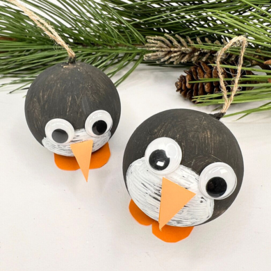 Zwei Anhänger aus bemalten Kugeln, die zu Pinguinen geschmückt wurden. Die Kugeln wurden schwarz und weiß angemalt und mit Wackelaugen sowie einem Schnabel und Füßen aus orangenen Fotokarton geschmückt.