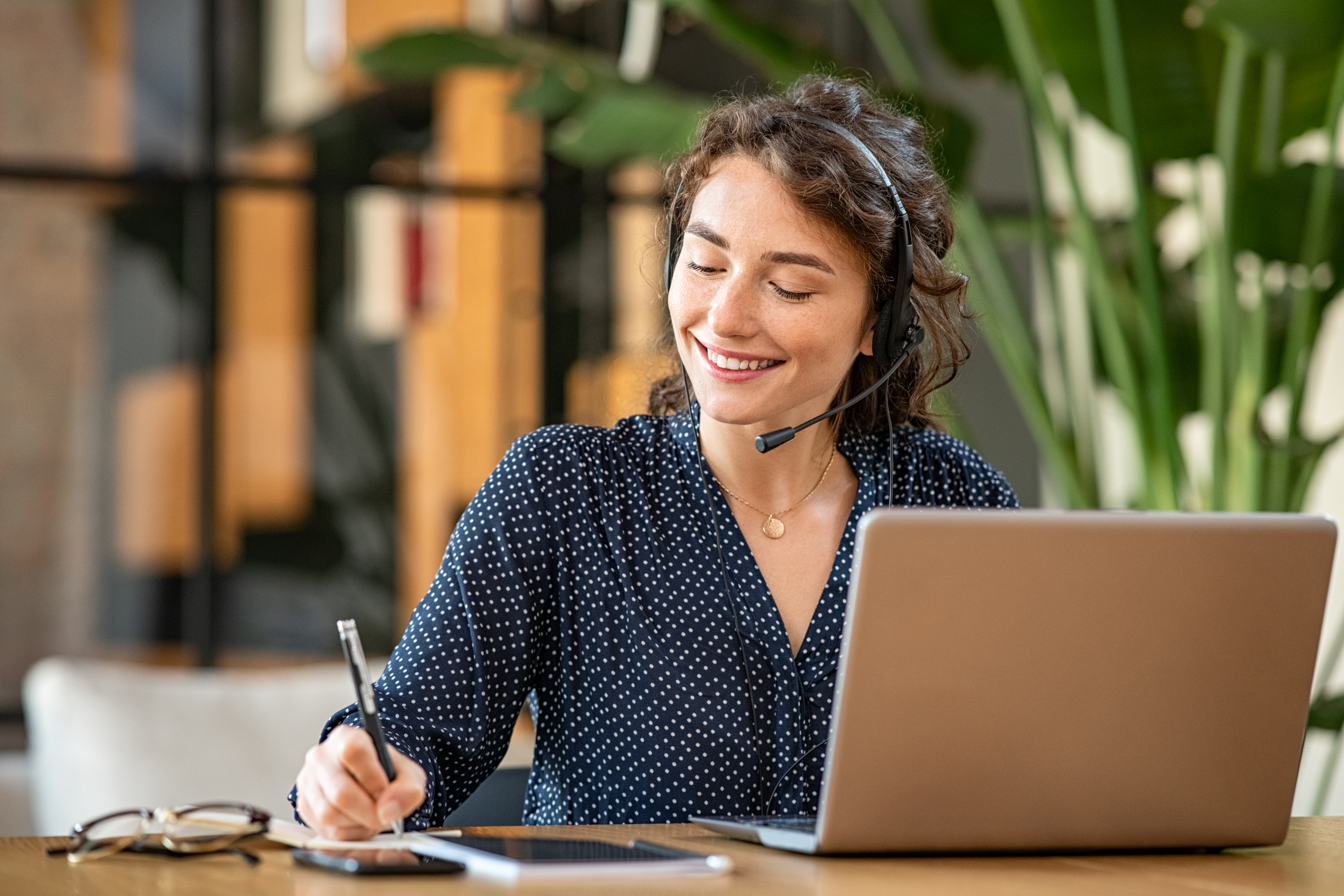 Lächelnde Frau mit Headset sitzt an einem Laptop und macht sich Notizen an einem Schreibtisch in heller Büroumgebung.