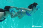 Zwei Personen tragen Schildkröt Junior Tauchmaske Tahiti Schwimmbrillen während sie sich an den Händen halten und in einem Pool unter Wasser schwimmen. Die rechte Person trägt einen blauen Bikini die linke Person trägt einen schwarzen. Das Schildkröt-Logo 