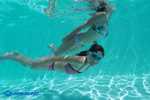 Zwei Mädchen schwimmen in Badeanzügen in einem Schwimmbad unter Wasser; eines der Kamera zugewandt trägt die Schildkröt Junior Schwimmbrille Bali 4+ blau mit Aufbewahrungstasche. Das Sonnenlicht mustert den Boden des Schwimmbeckens. Das SCHILDKRÖT-Logo ers