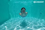 Ein Schwimmer trägt Schildkröt Junior Schwimmbrille rot (Bali 4+) unter Wasser in einem klaren blauen Pool. Das Sonnenlicht bildet wellenförmige Muster auf dem Beckenboden. Das Schildkröt-Logo ist in der oberen rechten Ecke zu sehen.