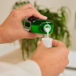 Jemand gießt eine weiße Flüssigkeit aus einer grünen Weleda Edeltannen-Erholungsbad 200 ml Flasche in einen kleinen Plastikbecher wobei eine Blattpflanze im Hintergrund verschwimmt.