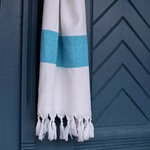 HängemattenGlück Strandtuch aus Bio-Baumwolle zweifarbig in 7 Farbkombinationen _31622 weiß-blau Das "Strandtuch aus Bio-Baumwolle" von HängemattenGlück in zweifarbigem Design hängt vor einer blauen Holztür mit Chevron-Muster. Das Handtuch ist in 7 Farbkombinationen erhältlich und verfügt über Fransen an den Enden. weiß-blau