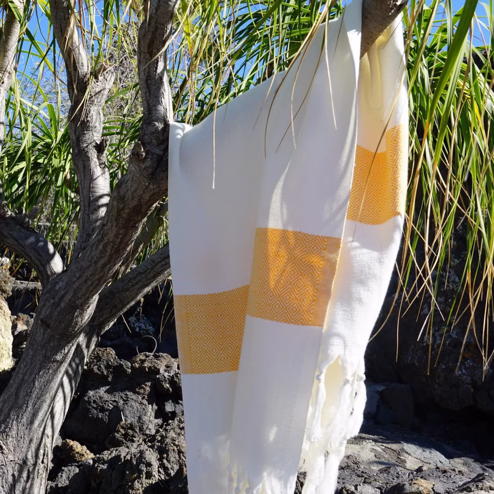 HängemattenGlück Strandtuch aus Bio-Baumwolle zweifarbig in 7 Farbkombinationen _31625 weiß-orange Das Strandtuch aus Bio-Baumwolle von HängemattenGlück hier in einer gelb-weißen Farbkombination hängt über einem Ast mit grünem Laub und steinigem Boden im Sonnenlicht. weiß-orange