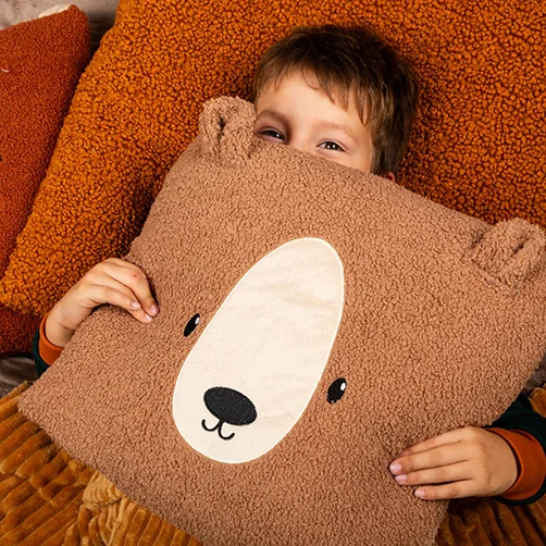 Ein Kind liegt gemütlich auf einer Couch und hält ein weiches Kissen in Bärenform vor das Gesicht. Die Umgebung ist mit warmen und kuscheligen Materialien dekoriert.