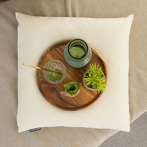 Eine weiße quadratische Kissenhülle, auf der ein runder Holztablett mit einem Glas, einer kleinen grünen Pflanze und einer dekorativen Flasche platziert ist. Das Bild vermittelt ein modernes und minimalistisches Wohngefühl.