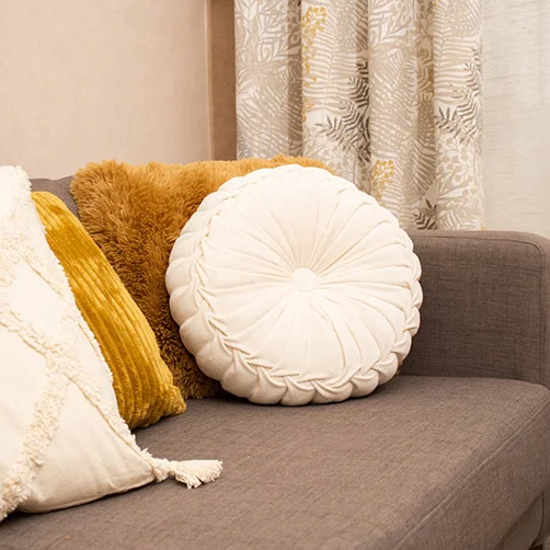 Ein rundes, cremefarbenes Kissen auf einem grauen Sofa, kombiniert mit weiteren dekorativen Kissen in Gelb- und Erdtönen. Im Hintergrund sind Vorhänge mit floralen Mustern zu sehen. Das Bild vermittelt Wärme und Gemütlichkeit.