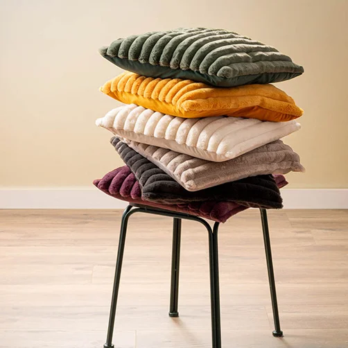 Ein Stapel aus Samtkissen in verschiedenen Farben (Grün, Gelb, Creme, Grau, Schwarz und Lila) auf einem modernen Metallhocker. Das Bild zeigt ein stilvolles und farbenfrohes Design.