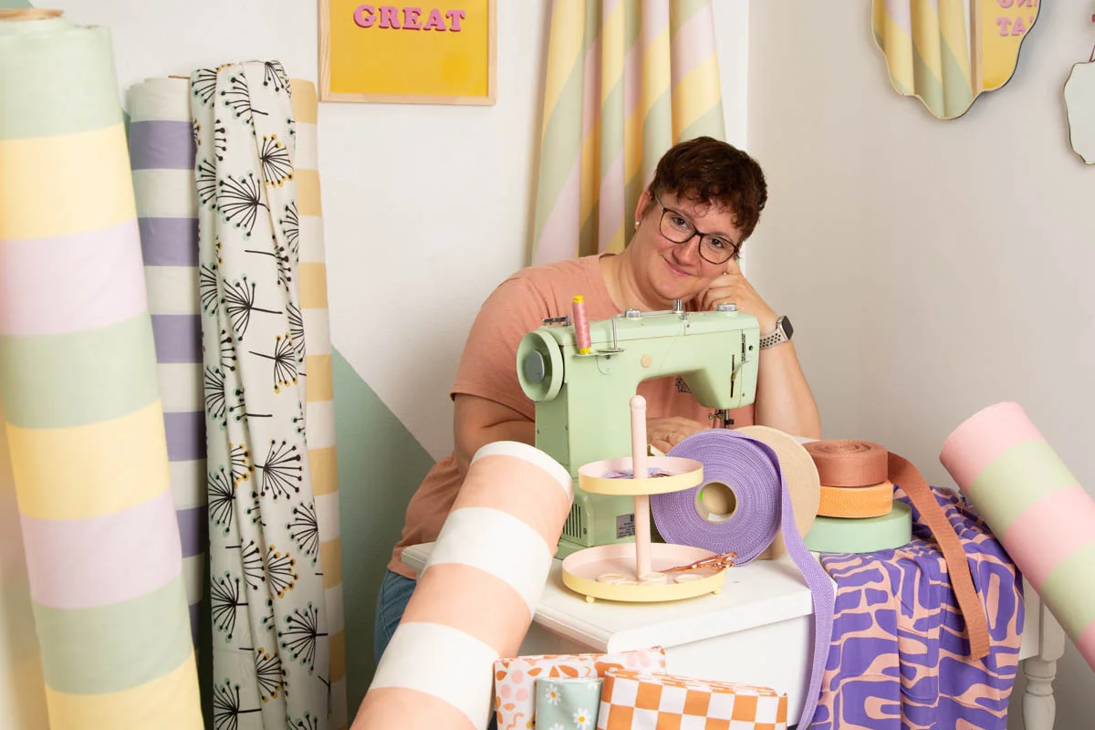 Person an Nähmaschine mit bunten Dekostoffen als Meterware in kreativem Arbeitsraum mit Motivationsschild