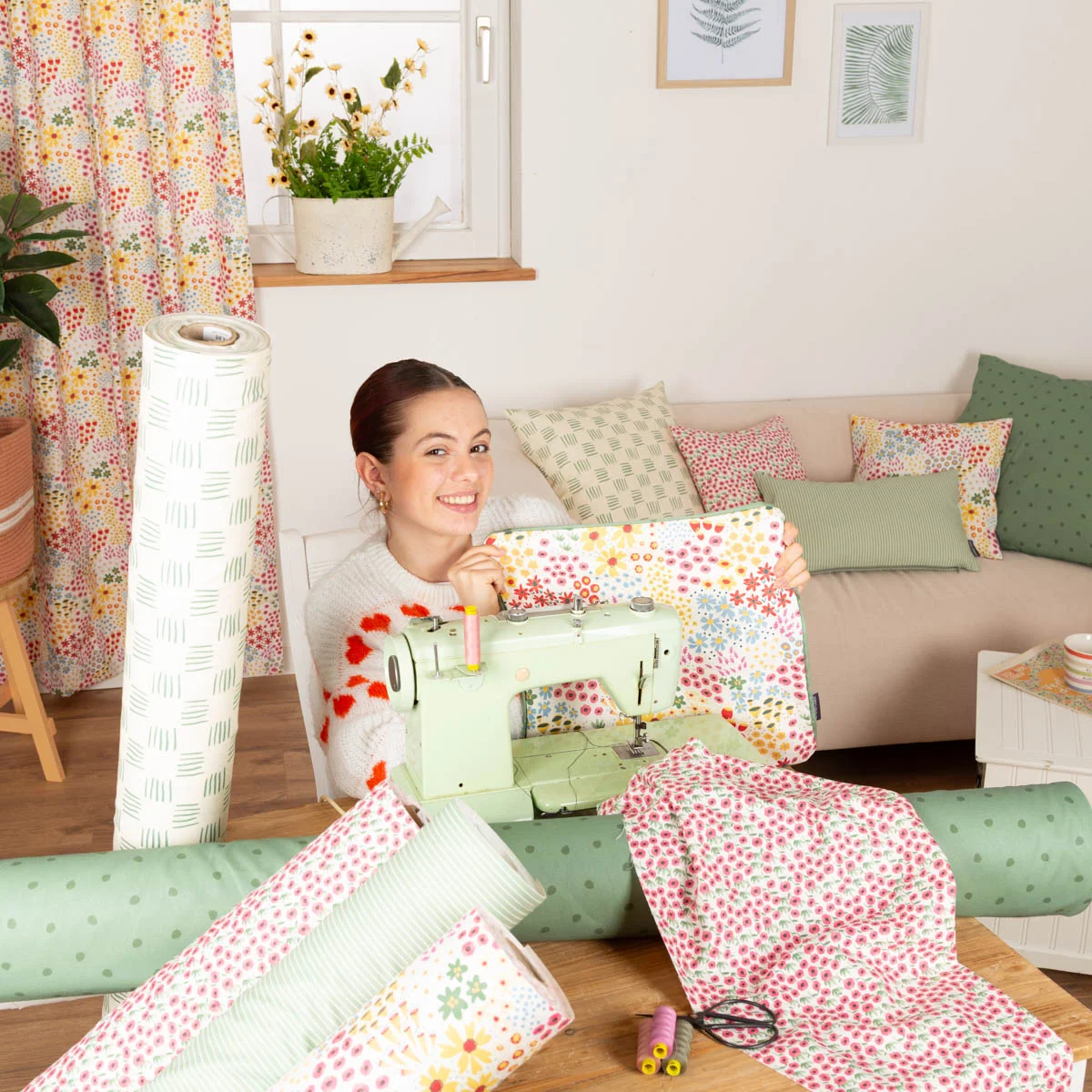 Lächelnde Frau präsentiert ein Nähprojekt aus fröhlichem Blumenstoff – umgeben von Stoffen als Meterware mit floralen Mustern, Punkten und Streifen in frischen Frühlingsfarben für kreative DIY-Ideen.