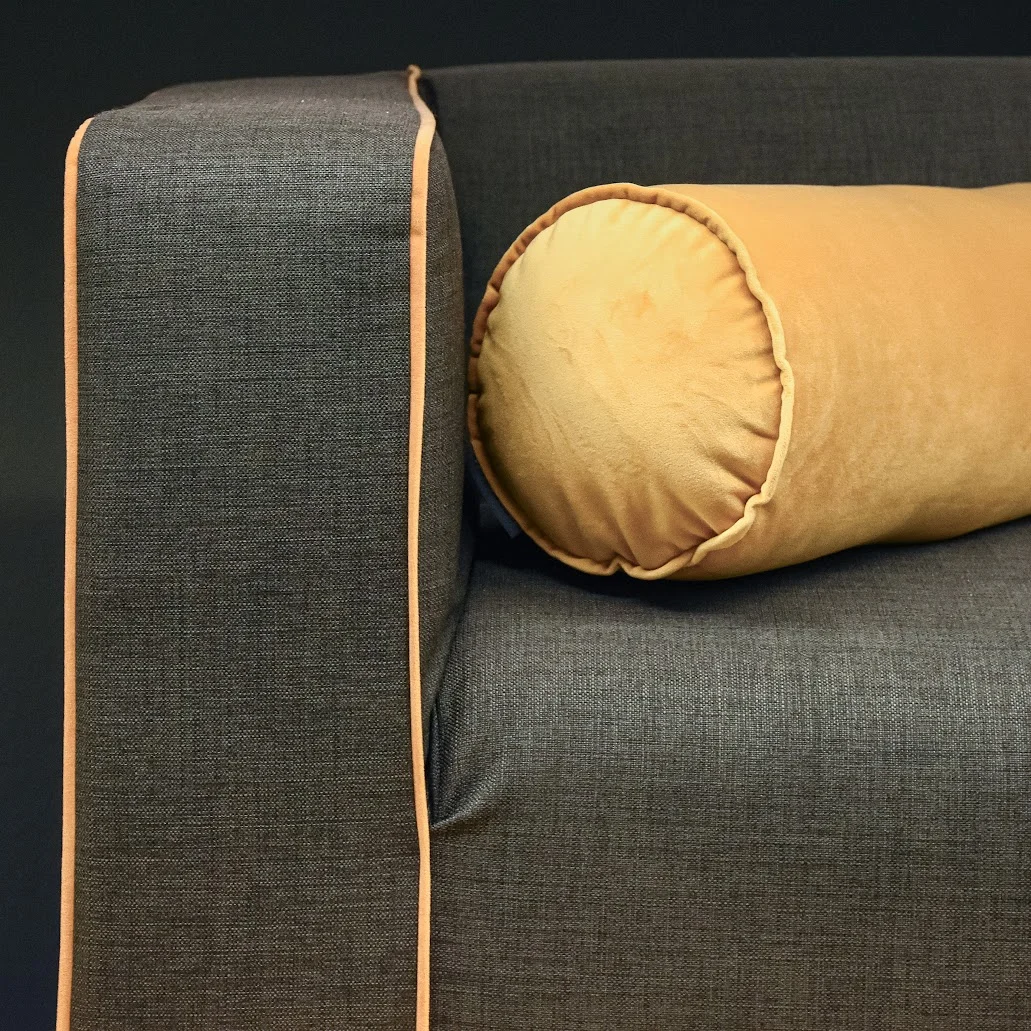 Detailansicht eines modernen Sofas mit dunkelgrauem Stoffbezug. Eine ockerfarbene Nackenrolle liegt dekorativ auf der Armlehne, die mit einer farblich abgestimmten Biese eingefasst
