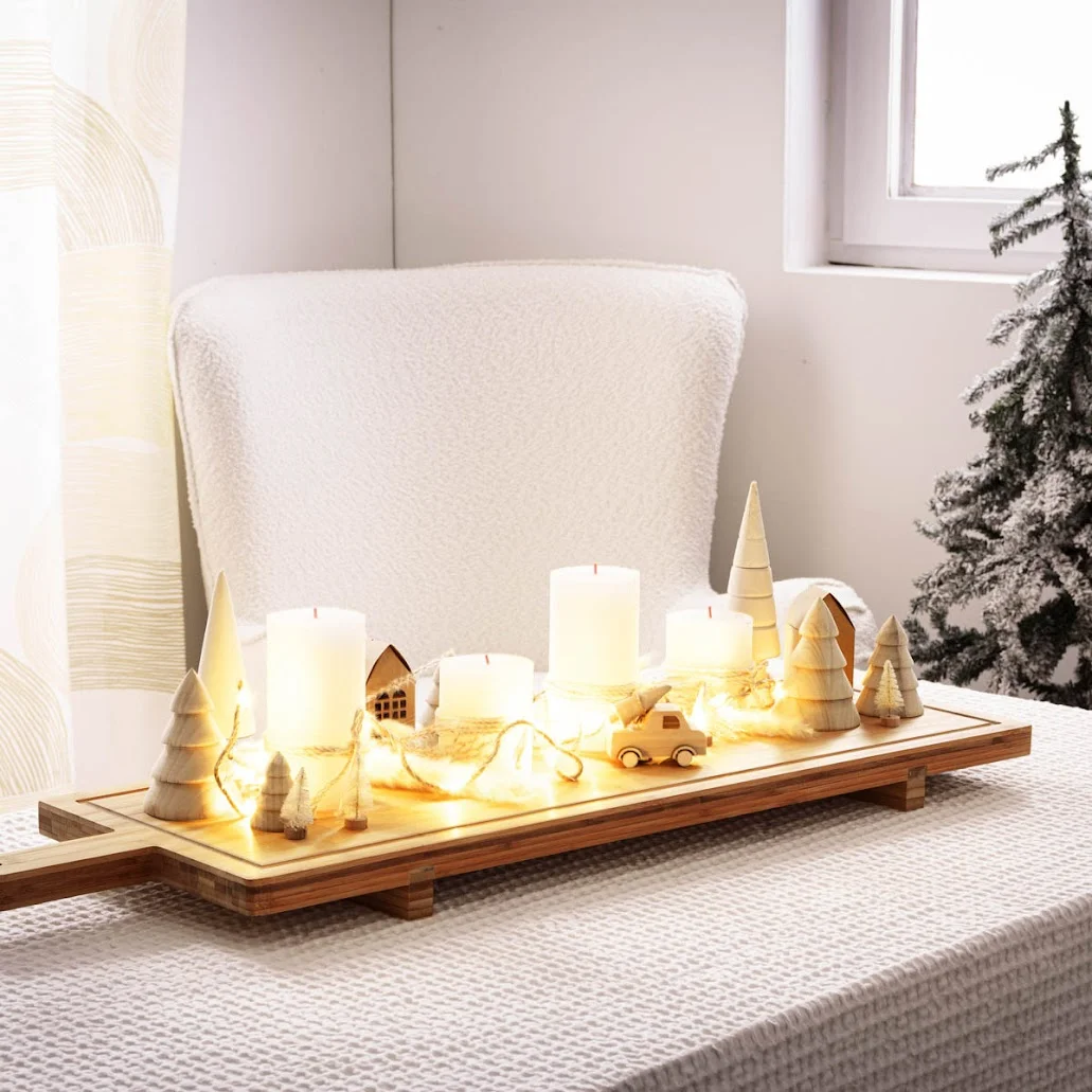 Weihnachtlich dekorierte Szene mit einem weißen Sessel und einer hellen Strukturdecke. Auf einem Tablett stehen vier weiße Kerzen, Holzfiguren wie Bäume und Häuschen sowie eine Lic