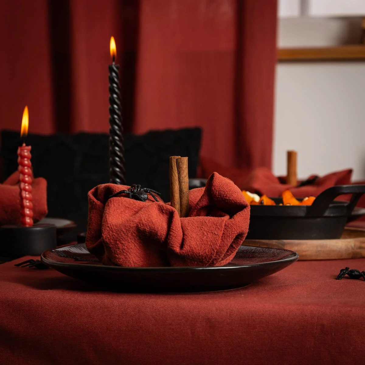 Detailaufnahme eines gedeckten Halloween-Tisches mit roter Tischwäsche, schwarzem Kerzenständer und roter Stoffserviette in Form eines Kürbisses mit Zimtstange als Stiel und einer kleinen Dekospinne.