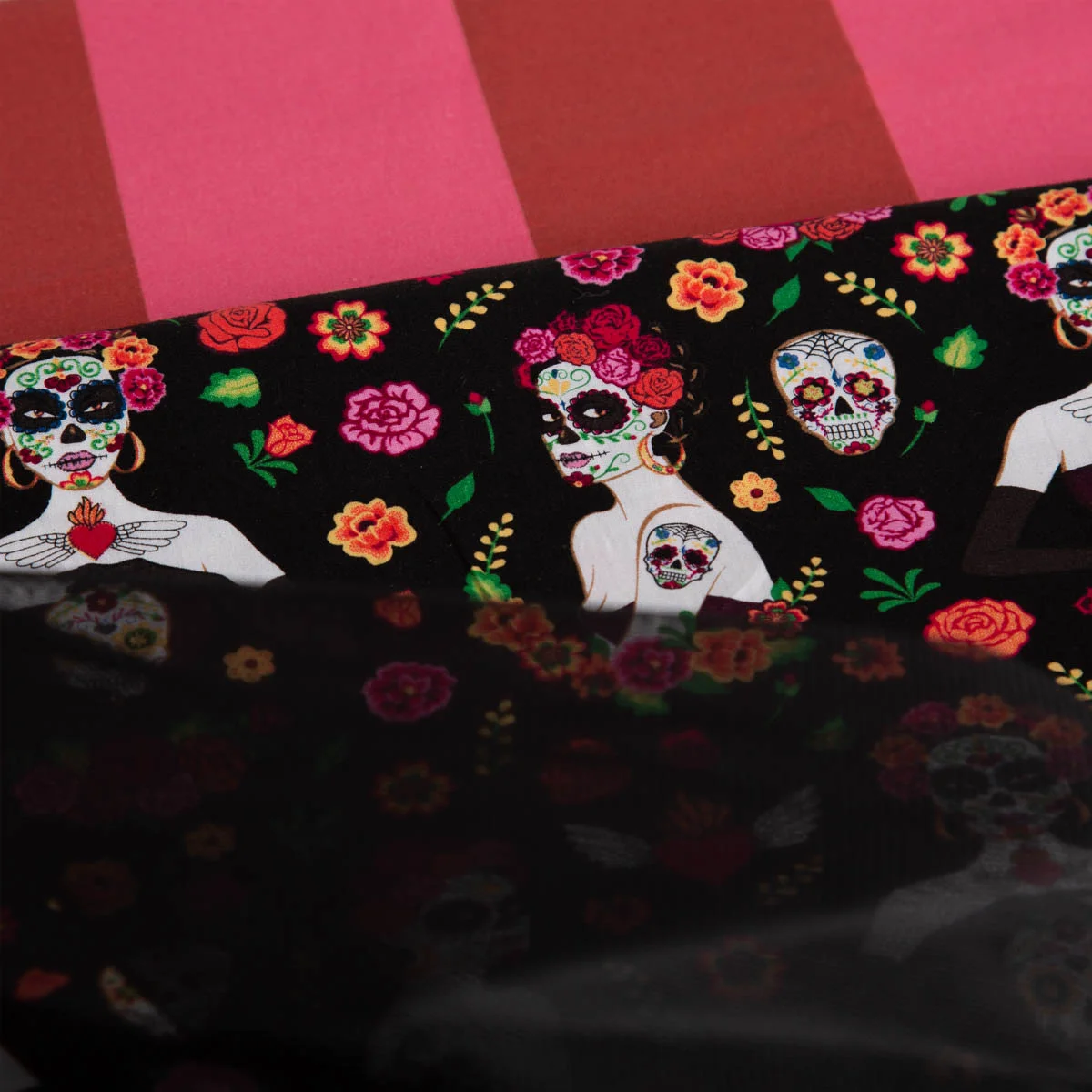 Stoff mit buntem „Día de los Muertos“-Motiv: Frauen mit Totenkopf-Make-up, bunte Totenschädel und Blumen auf schwarzem Hintergrund. Im Hintergrund ein pink-rot gestreifter Stoff.