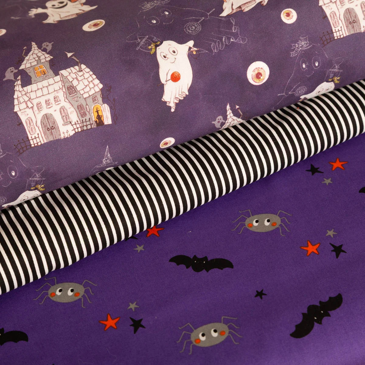 Drei Stoffrollen mit Halloween-Motiven: oben violetter Stoff mit Geistern und Spukhäusern, in der Mitte schwarz-weiß gestreifter Stoff, unten violetter Stoff mit Spinnen, Fledermäusen und Sternen.