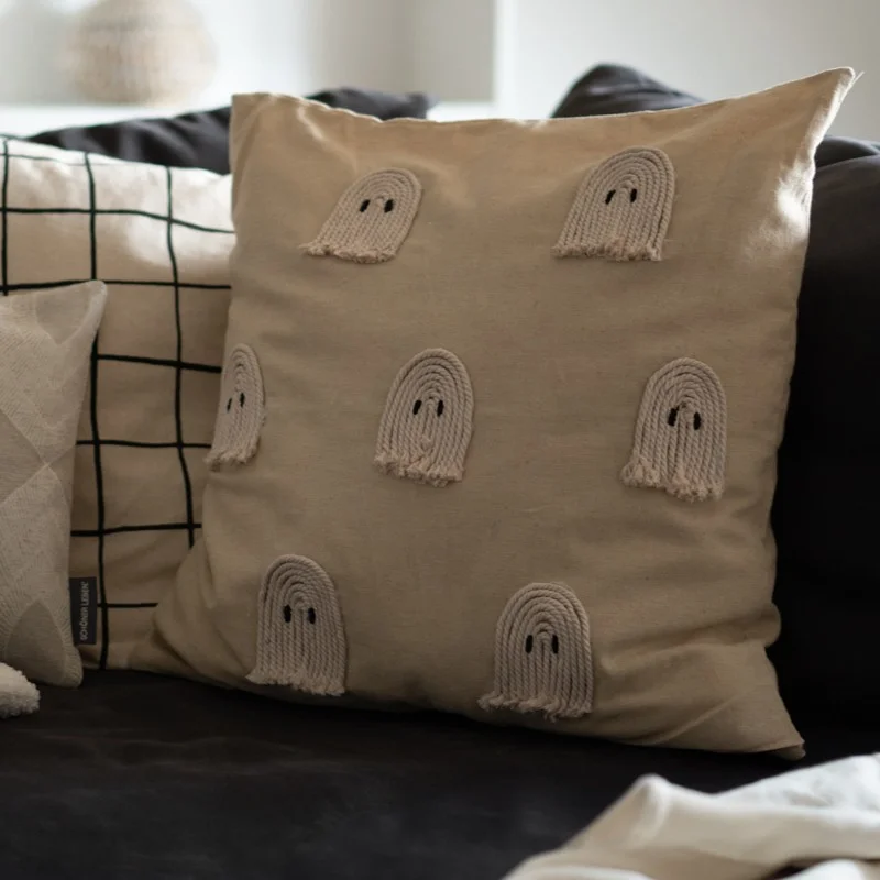 Zierkissen auf einem Sofa, eines davon mit applizierten kleinen Geistern aus hellem Seil auf naturfarbenem Bezug. Dahinter weitere Kissen mit Karomuster und geometrischen Designs.