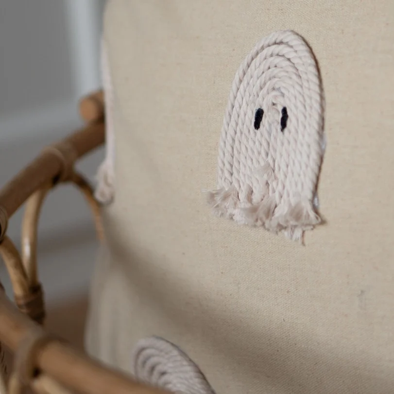 Nahaufnahme eines Kissens mit einem applizierten kleinen Geist aus geflochtener Baumwollkordel auf naturfarbenem Stoff, platziert auf einem Rattanstuhl.