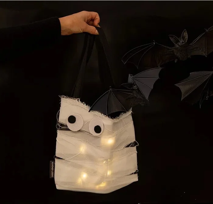 Hand hält eine selbstgebastelte Halloween-Tasche im Mumien-Look mit aufgeklebten Augen, Lichterkette im Inneren und schwarzen Tragegriffen. Im Hintergrund fliegende Fledermäuse auf schwarzer Wand.