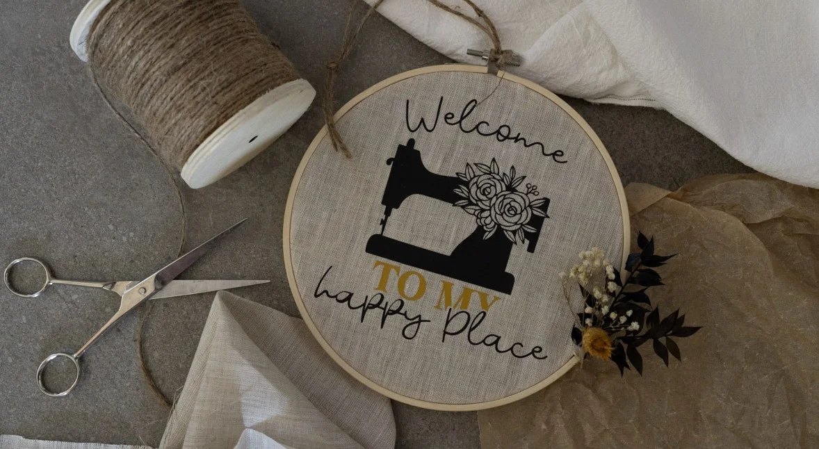 Ein bestickter Stickrahmen mit Nähmaschinenmotiv und floralen Details, begleitet von Schere und Garn – symbolisch für Bastelbedarf und Nähzubehör als „Happy Place“