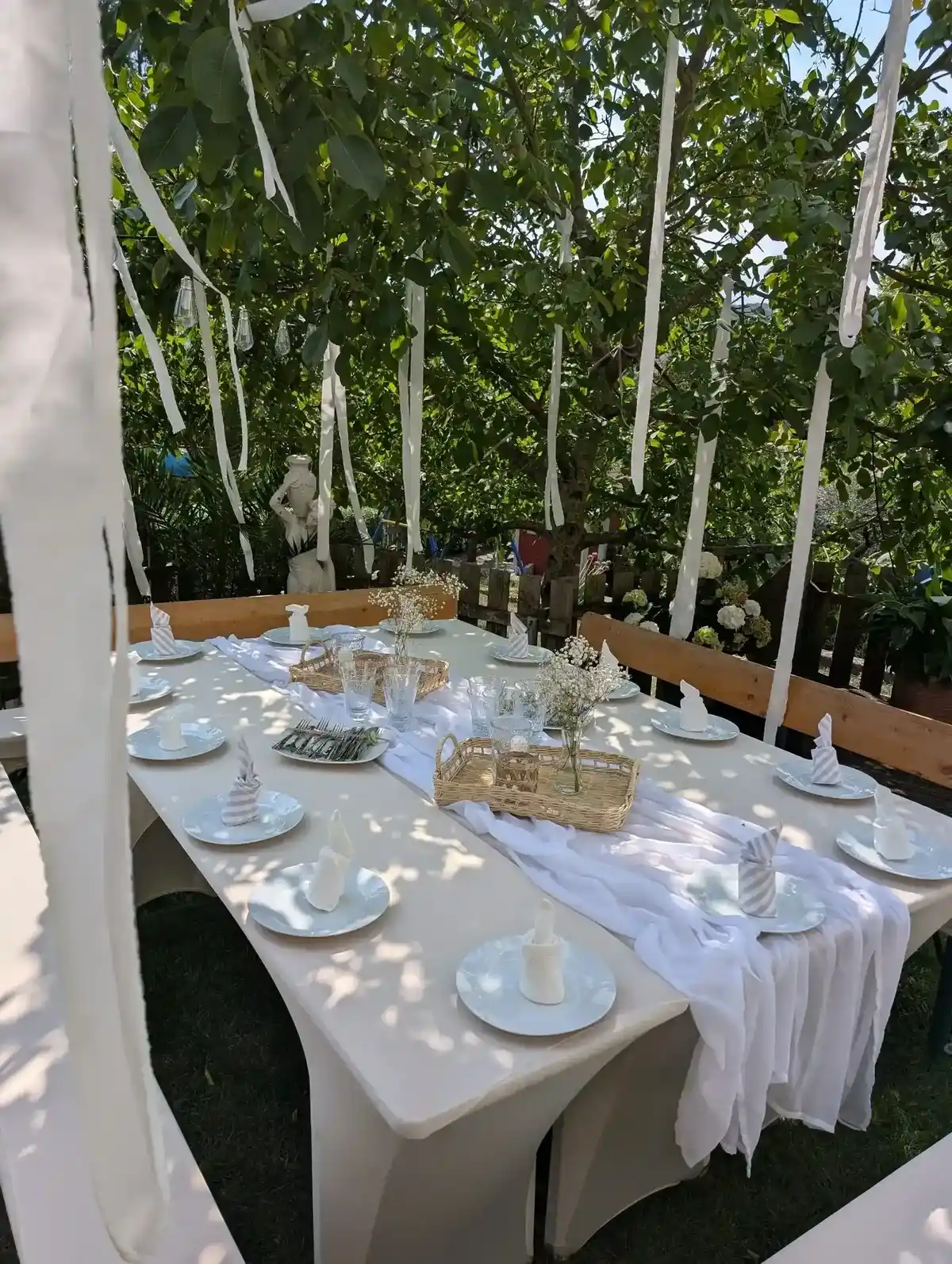 Gartenparty mit weißer Deko – lange Tafel mit Stoffstreifen in den Bäumen, Stoffläufer und Servietten.