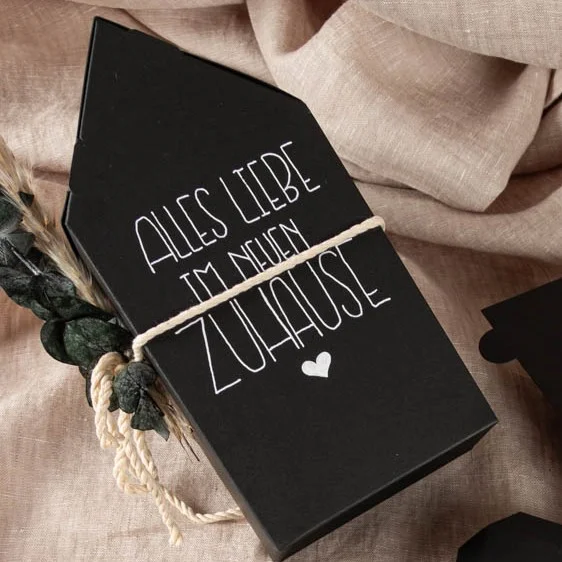 Schwarze Geschenkverpackung in Hausform auf beigem Stoff. Darauf steht in weißer Schrift „Alles Liebe im neuen Zuhause“, verziert mit einem kleinen Herz und umwickelt mit einer naturfarbenen Kordel.