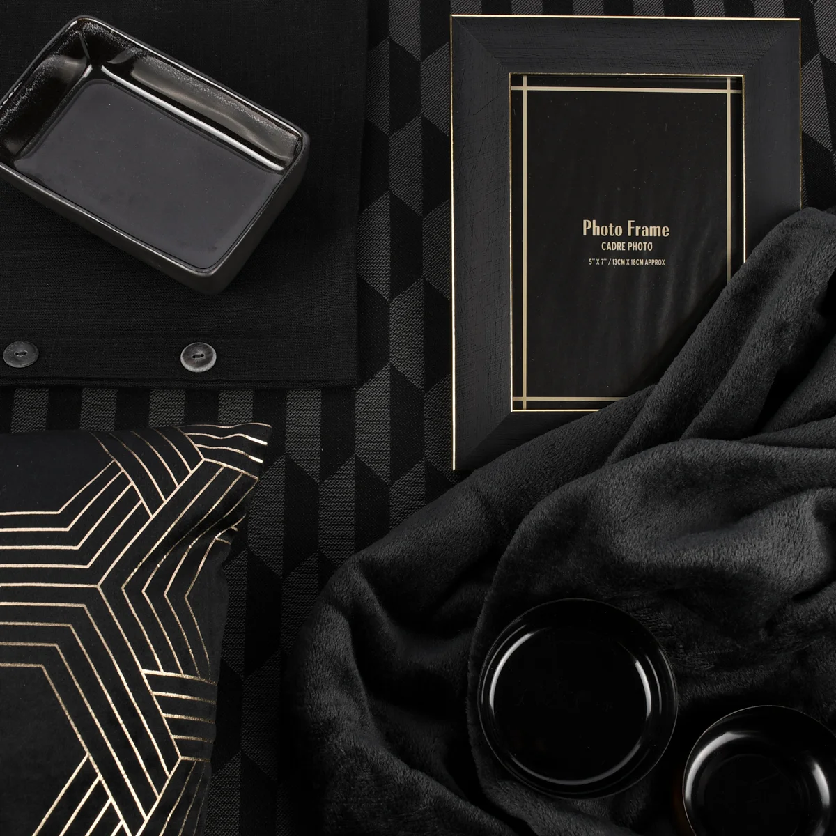Flatlay in Schwarz-Gold mit geometrischem Muster: Foto­rahmen, schwarze Schale, Kissen mit goldenen Linien, weiche Decke und kleine Accessoires wie Tassen auf schwarzem Hintergrund mit Würfelmuster.