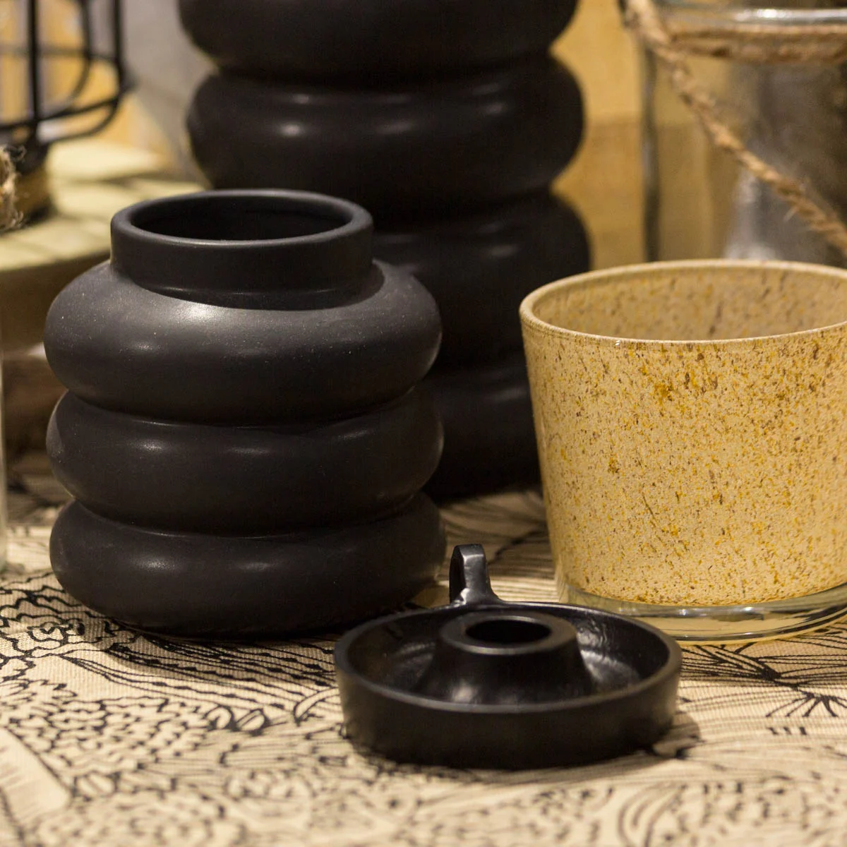 Stillleben mit schwarzer, ringförmiger Keramikvase, beigefarbenem Becher mit Sprenkeln und flachem schwarzen Kerzenhalter. Die Deko steht auf einem Tisch mit gemustertem Stoff in Naturtönen.