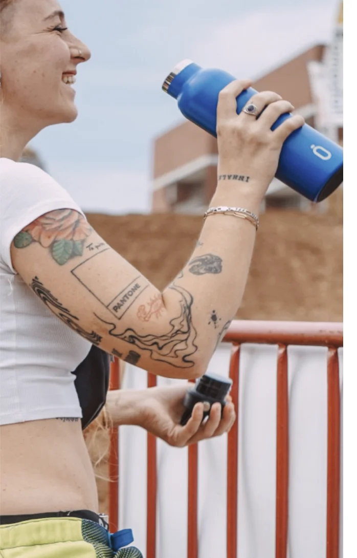 Junge Frau mit Tattoos trinkt unterwegs aus einer blauen Runbott Trinkflasche – stylische Geschenkidee für aktive Menschen