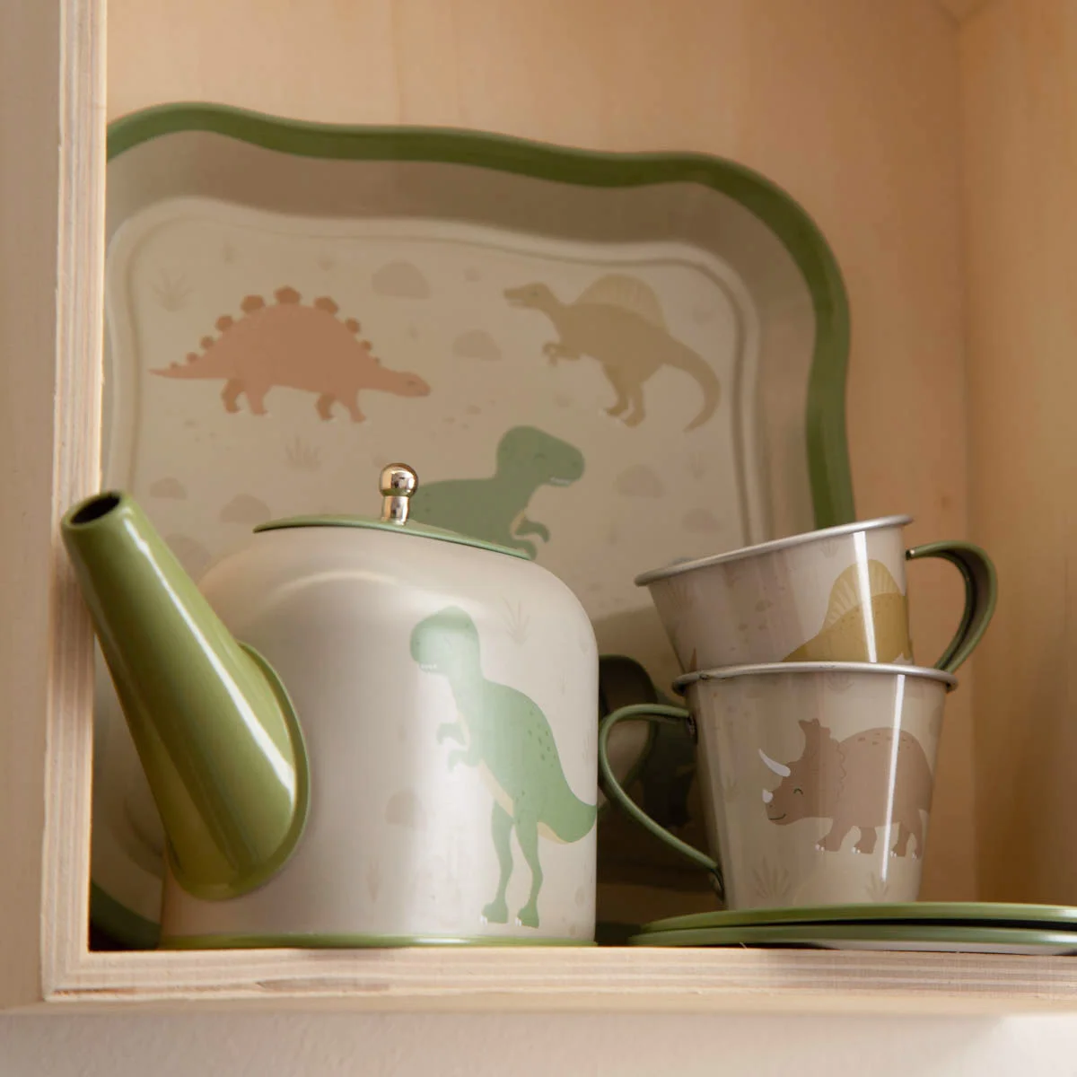 Weiß-grüner Teekessel und Tassen mit bunten Dinosauriermotiven, dahinter ein Tablett mit passenden Dino-Illustrationen.