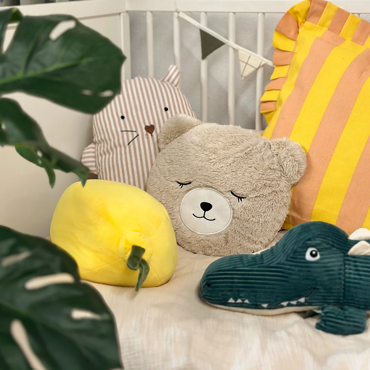 Verschiedene Kuschelkissen in Tier- und Obstform, darunter ein Bär, eine Katze, ein Krokodil und eine gelbe Zitrone, dekoriert mit bunten Kissen im Kinderbett.