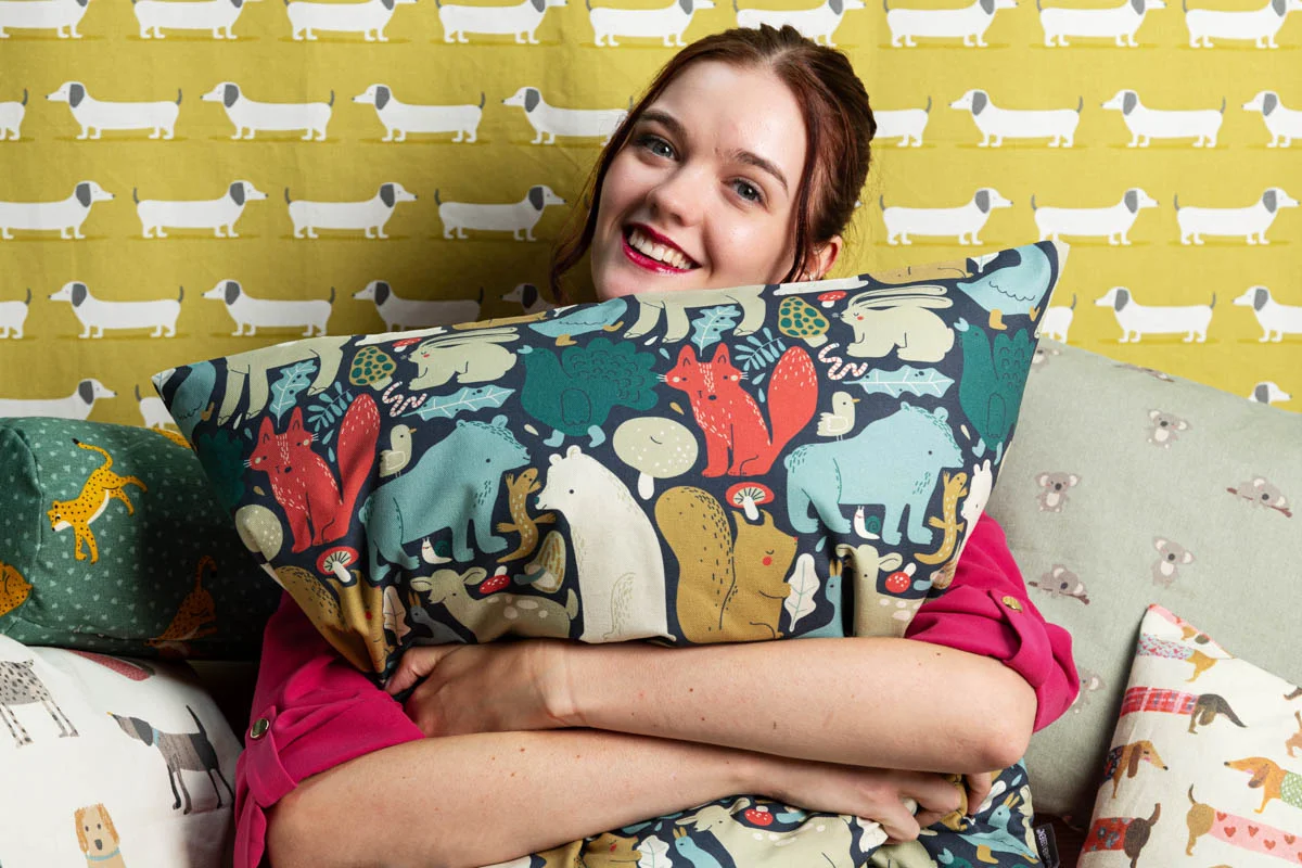 Eine lächelnde Frau hält ein buntes Kinderzimmerkissen mit Tiermotiven, darunter Fuchs, Bär und Hase, in den Armen. Im Hintergrund sind weitere Kissen und Stoffe mit Hundemotiven und kindgerechten Designs zu sehen – handgefertigte Produkte aus hochwertigen Stoffen von SCHÖNER LEBEN, ideal für individuelle Kinderzimmergestaltung.