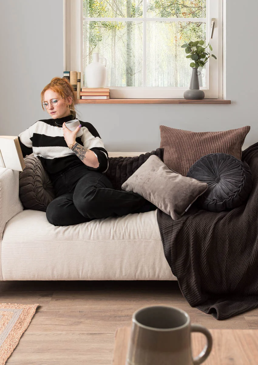 Frau sitzt auf dem Sofa zwischen grauen Kissen und liest ein Buch